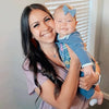Woman holding a baby with a blue bow in a home setting wearing necklace