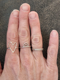Hand wearing three rings one with a heart design on a dark surface