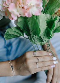 Hand holding a bouquet of pink flowers with green leaves, wearing rings and a bracelet.